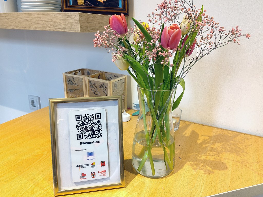 Die Plakette mit dem QR-Code, wie sie auch am Gedenkstein steht, mit einem Strauß Blumen. Foto. Hensel