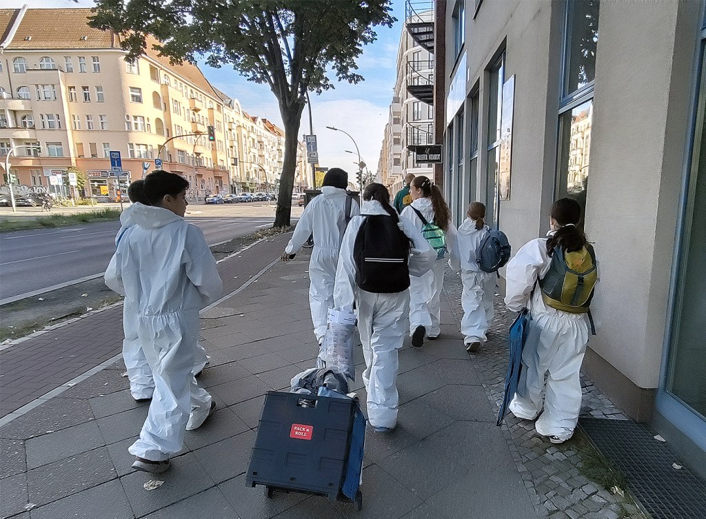 Gruppe von Schulkindern in weißen Schutzanzügen und mit Rucksäcken läuft auf einem Gehweg im Wedding.