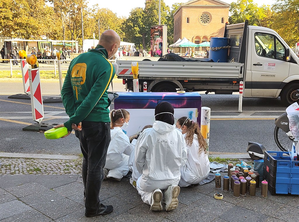 Kinder in weißen Schutzanzügen arbeiten an einem Stromkasten, daneben stehen zahlreiche bunte Sprühdosen und ein Betreuer.